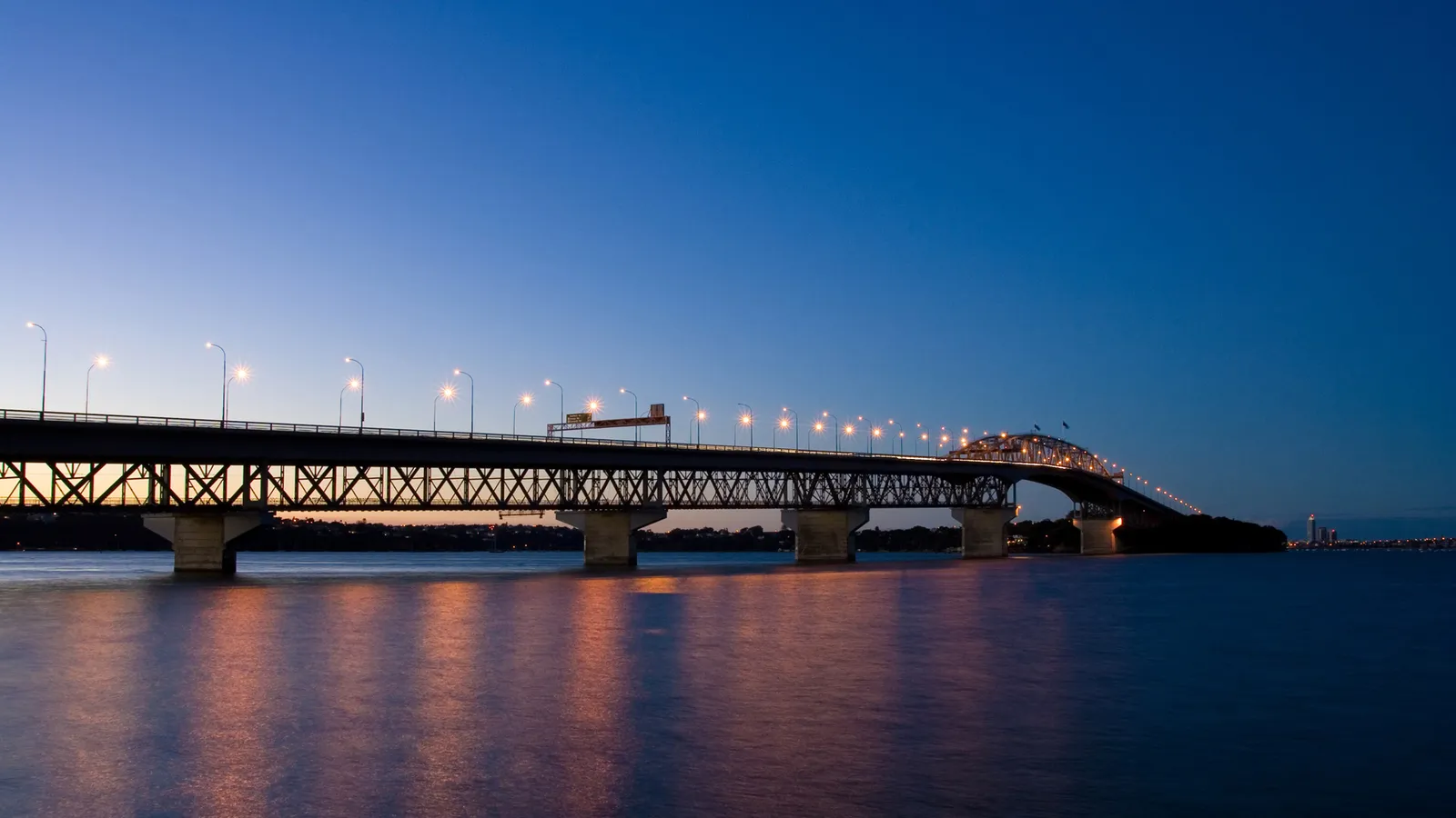 A long bridge spanning across a body of water at dusk, with lights illuminating the roadway and reflections on the water.