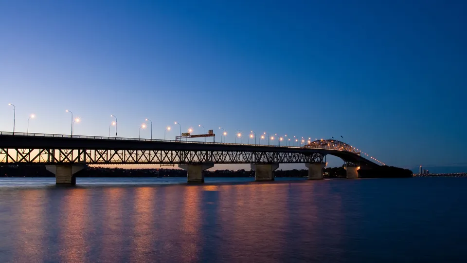 A long bridge spanning across a body of water at dusk, with lights illuminating the roadway and reflections on the water.