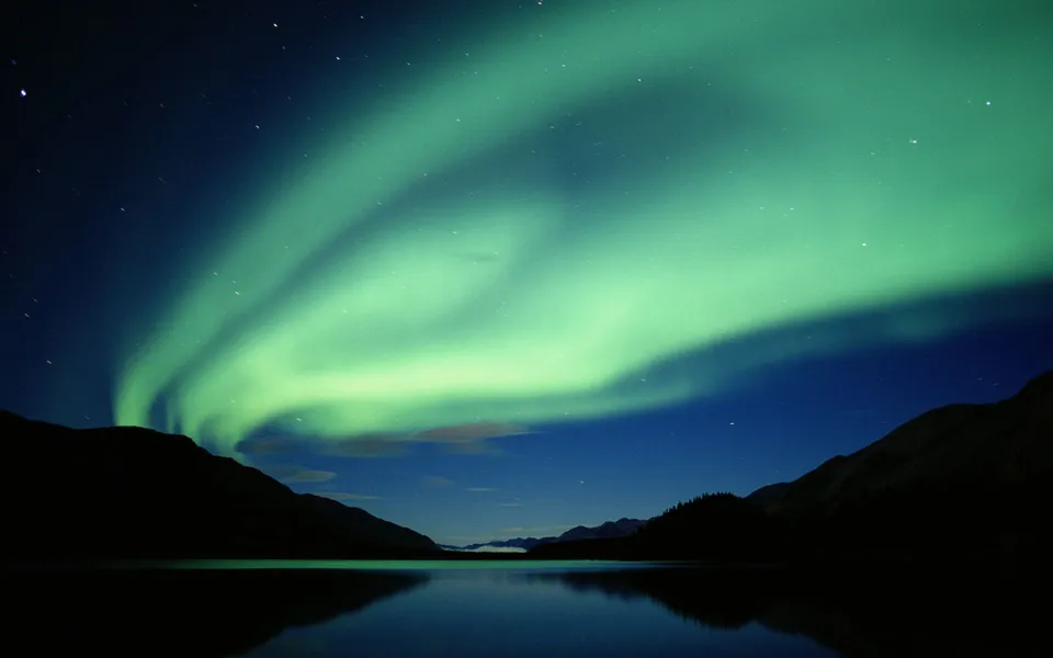 The green aurora borealis glows in the night sky above a dark mountain landscape and a still lake.