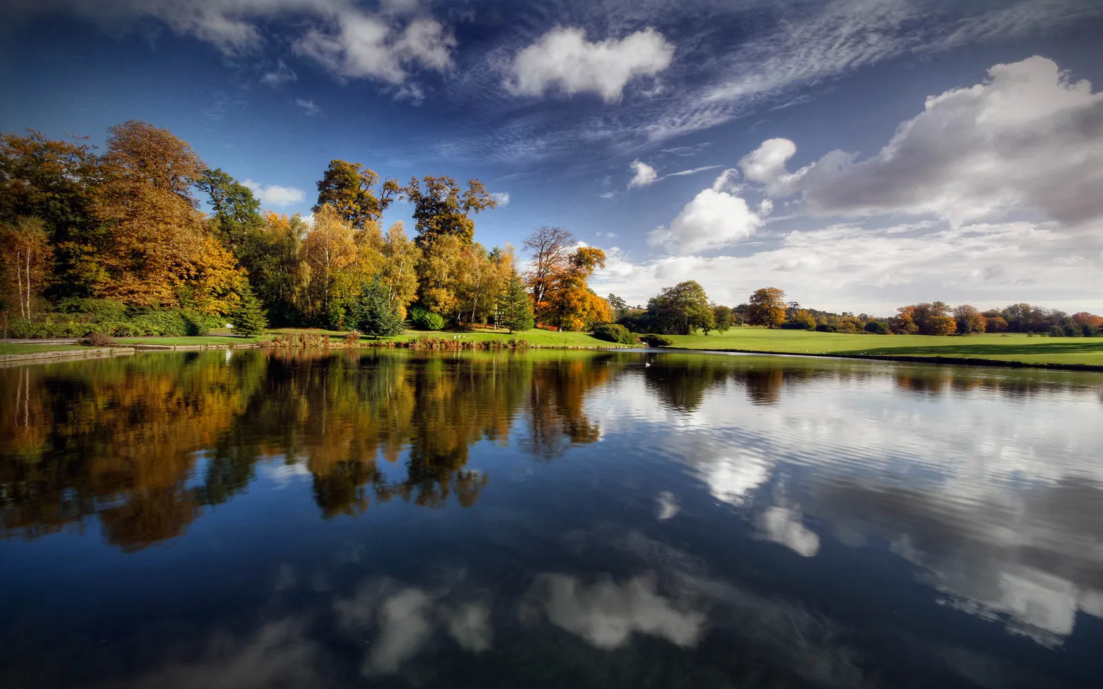 A tranquil lake reflects colorful autumn trees and a dramatic sky with clouds.