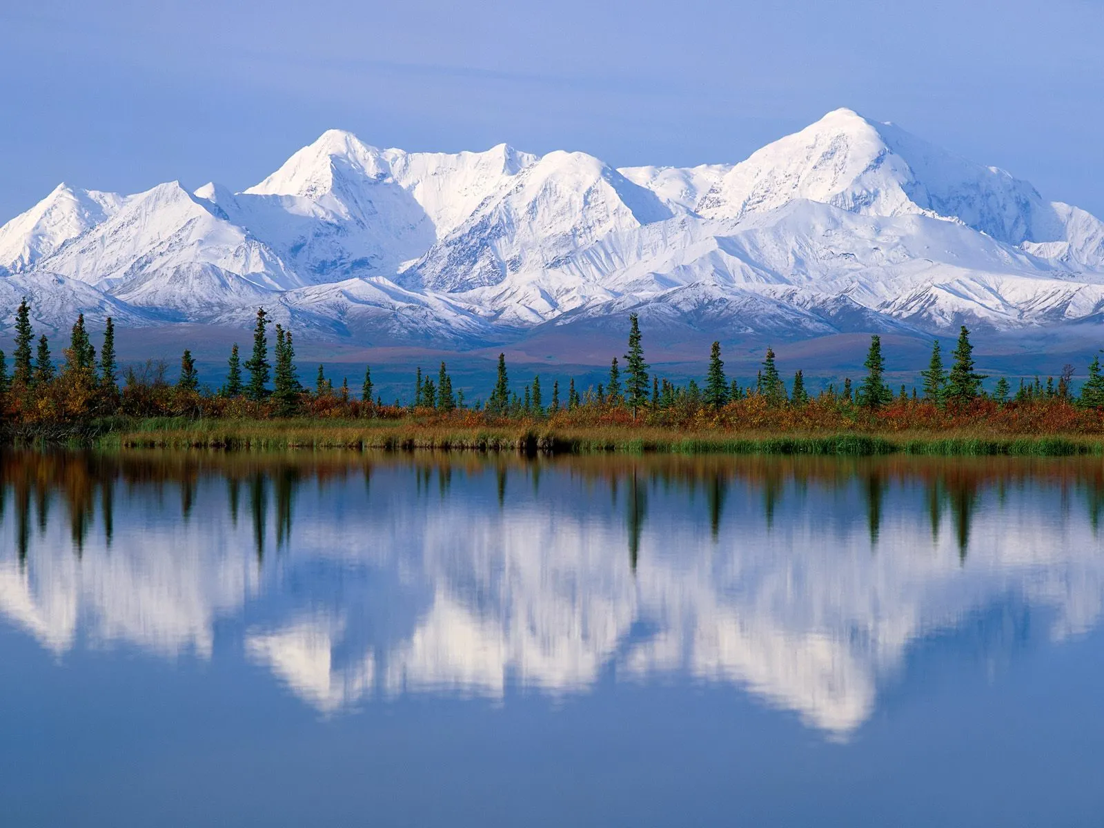 Snow-covered mountains reflect in a calm lake with autumn foliage.