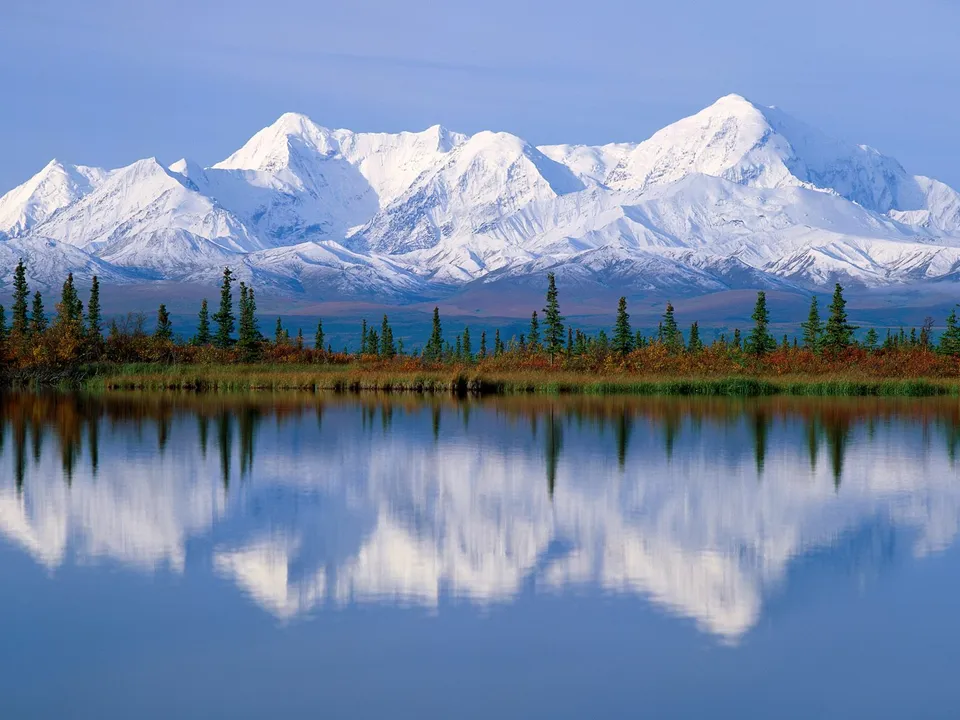 Snow-covered mountains reflect in a calm lake with autumn foliage.