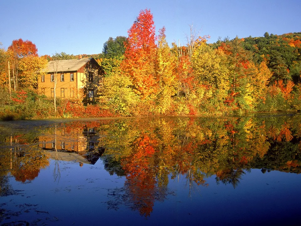A yellow house sits beside a lake surrounded by trees in peak autumn foliage, with reflections in the water.