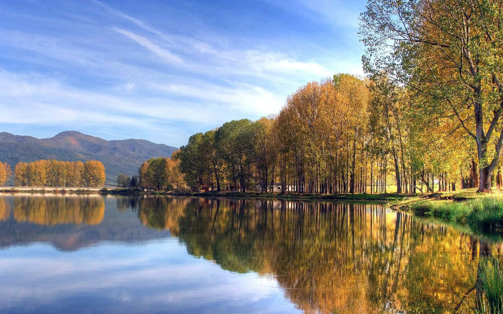 A calm lake reflects trees with autumn foliage and distant mountains under a blue sky with wispy clouds.