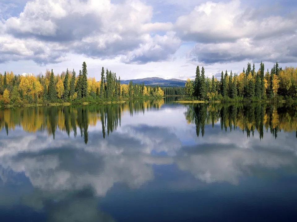A calm lake perfectly reflects a shoreline lined with autumn trees and a cloudy sky.
