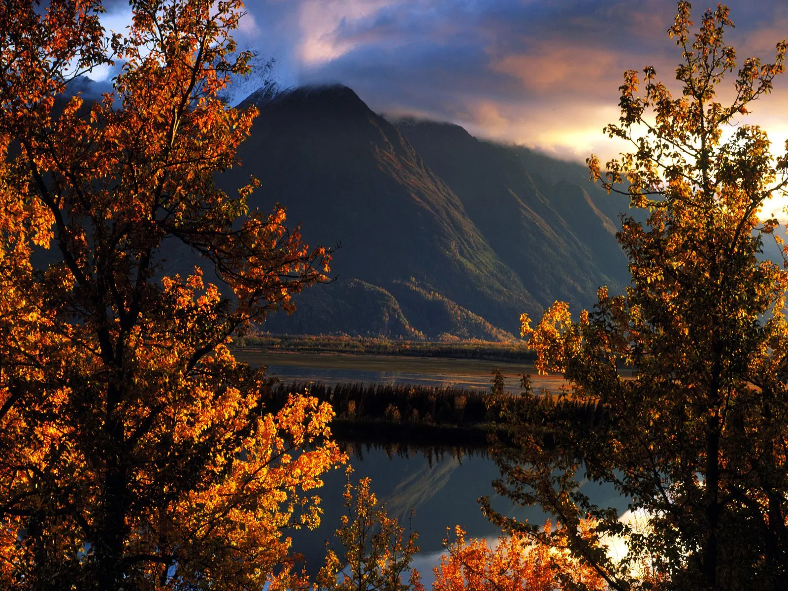 Framed by vibrant autumn trees, a calm lake reflects a majestic mountain under a dramatic sky at sunset.