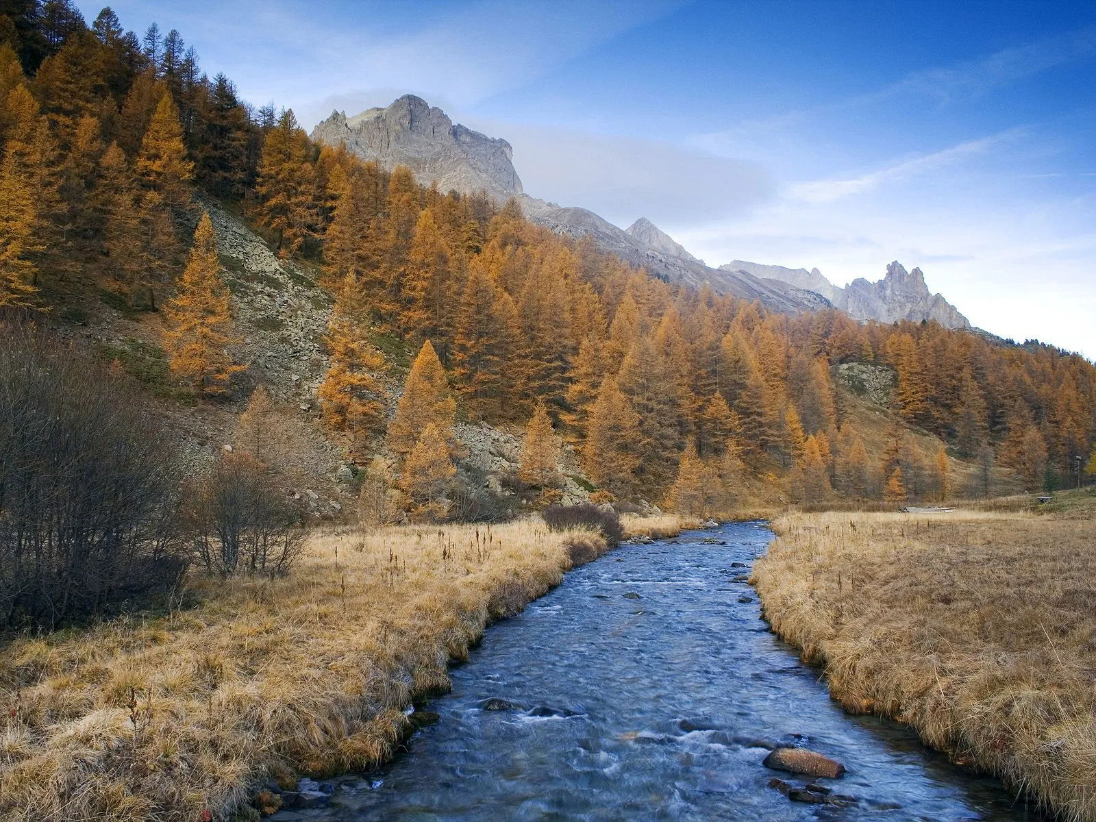 A clear blue river flows through a valley with golden autumn trees and rocky mountains in the background.