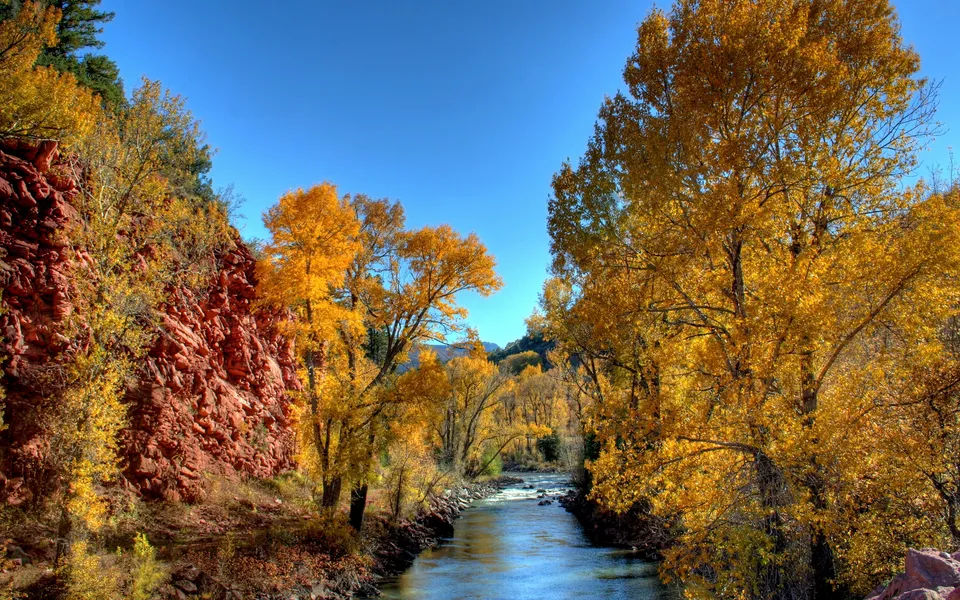 A river flows through a landscape with vibrant golden autumn trees and red rock formations under a clear blue sky.