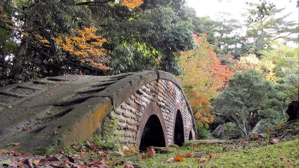 An old stone bridge with two arches covered in moss and lichen, surrounded by trees displaying autumn colors.