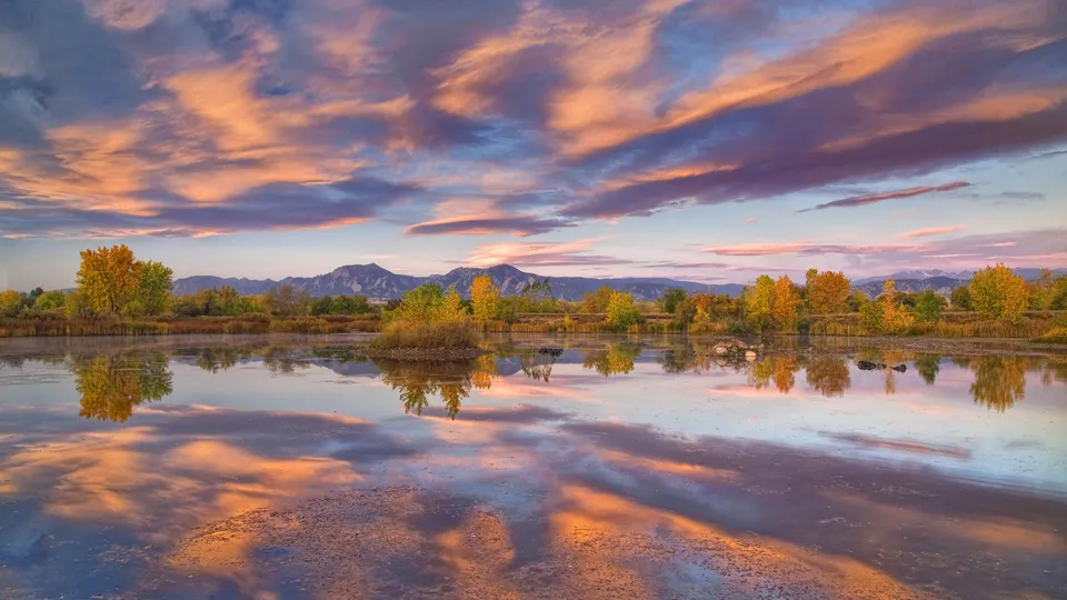 A serene lake reflects the vibrant colors of an autumn sunset sky and distant mountains.