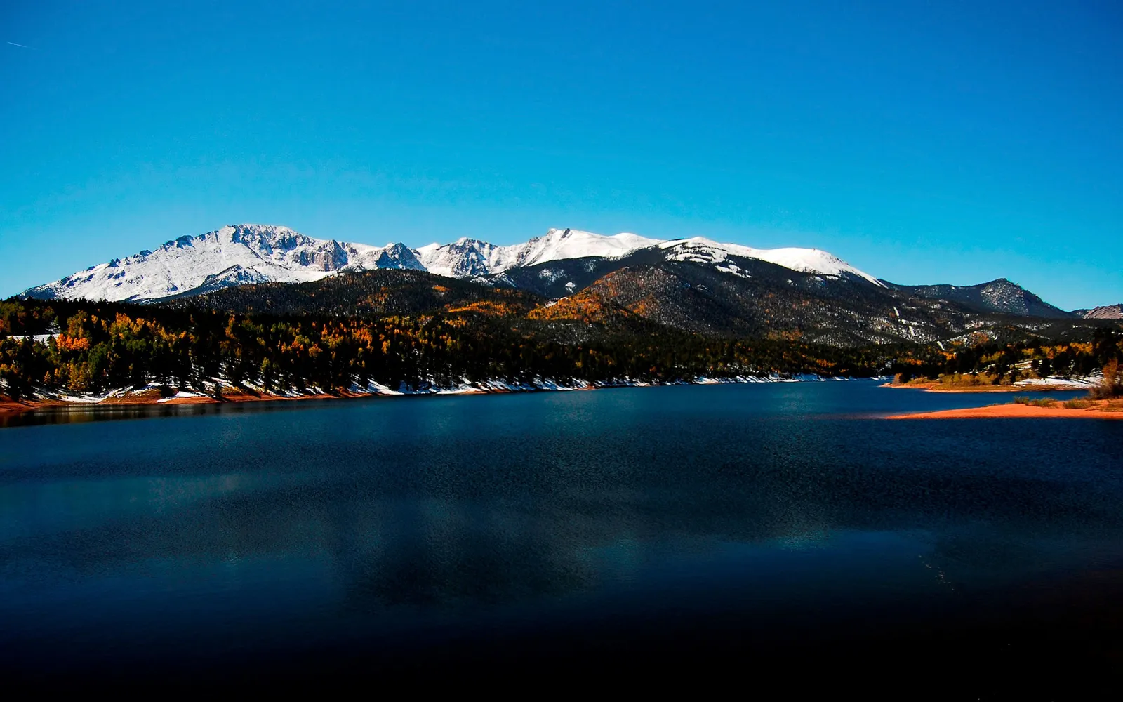 Snowy mountain peaks contrast with autumn trees reflected in a dark lake.