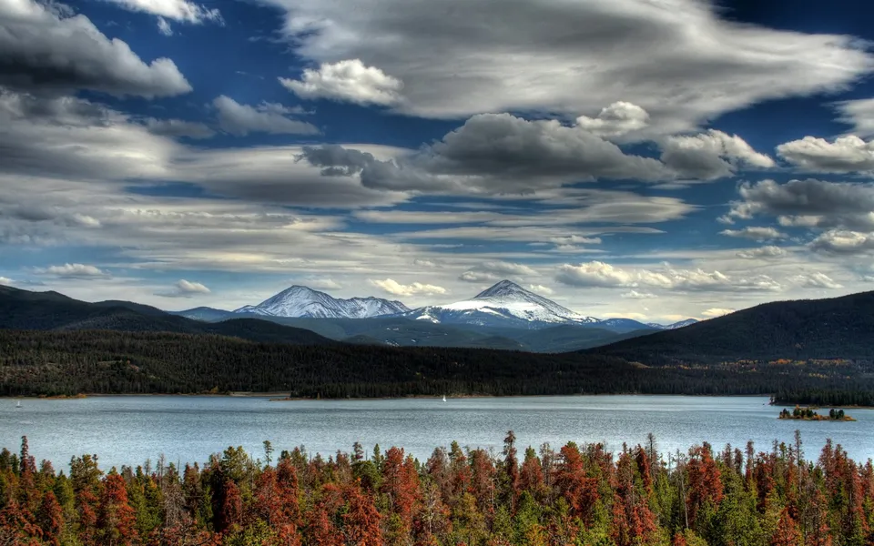 Snow-capped mountains loom over a lake, with foreground trees showing autumn colors.