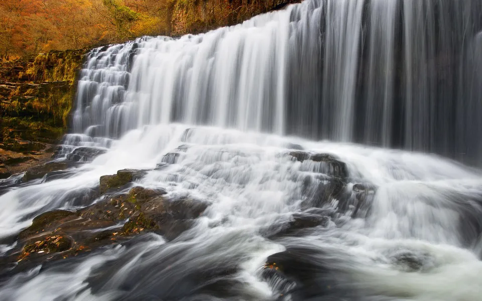 A wide, multi-tiered waterfall flows over dark rocks, with autumn foliage visible on the left bank.