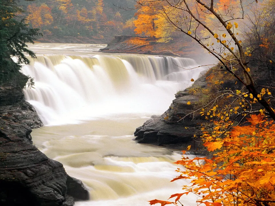A wide waterfall flows over rocks, surrounded by trees with vibrant orange and yellow autumn leaves.