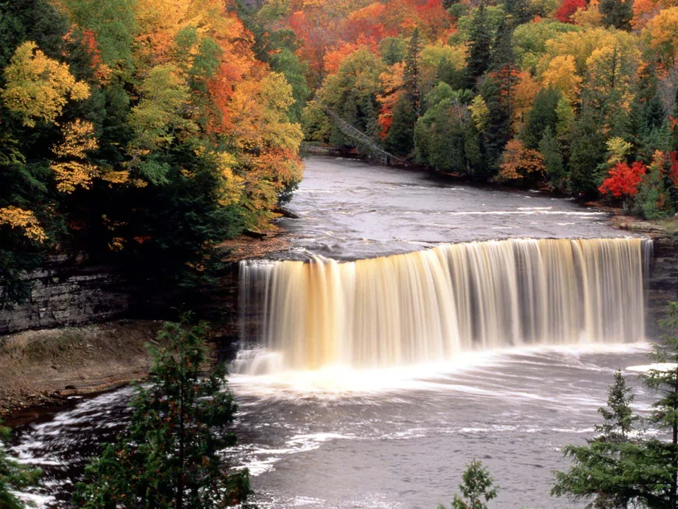 A wide waterfall cascades over a rocky ledge, surrounded by trees displaying brilliant autumn colors.