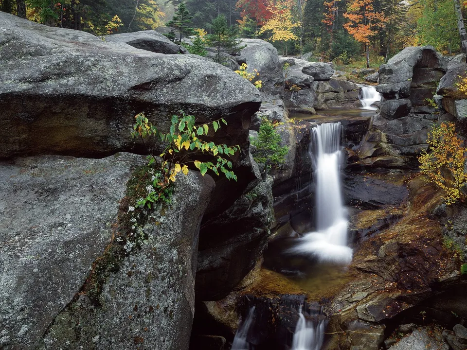 A multi-tiered waterfall cascades over dark, moss-covered rocks surrounded by autumn foliage.