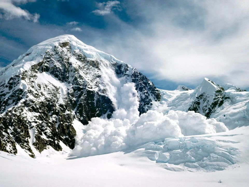 A large avalanche is actively cascading down a steep, snow-covered mountain face under a blue sky with clouds.