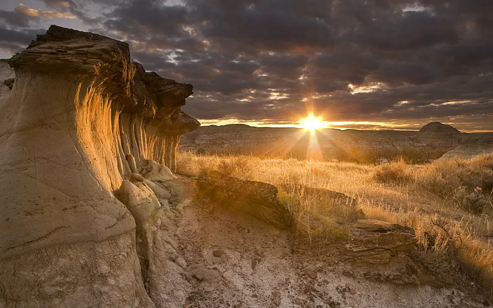 Sunlight streams through dramatic clouds over eroded rock formations and dry grass in a desert landscape.