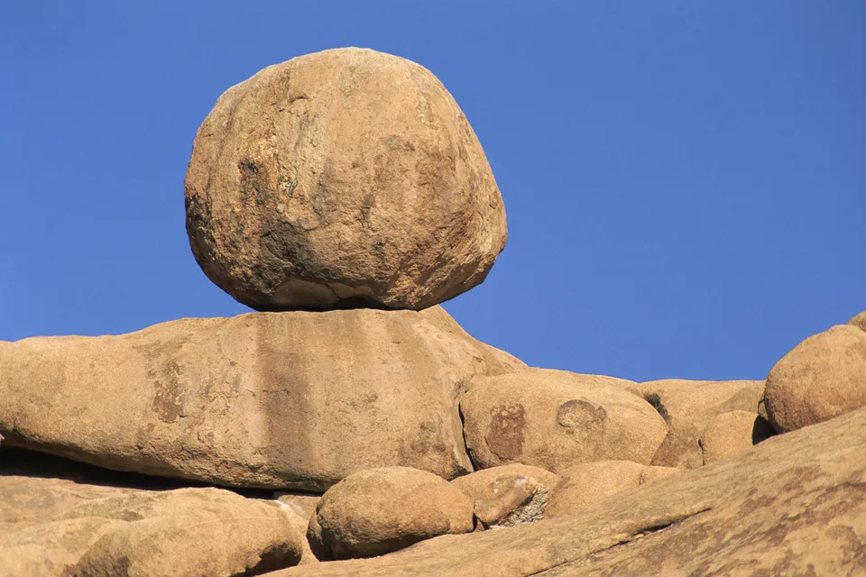 A large, round boulder is balanced on top of a flat rock formation against a clear blue sky.
