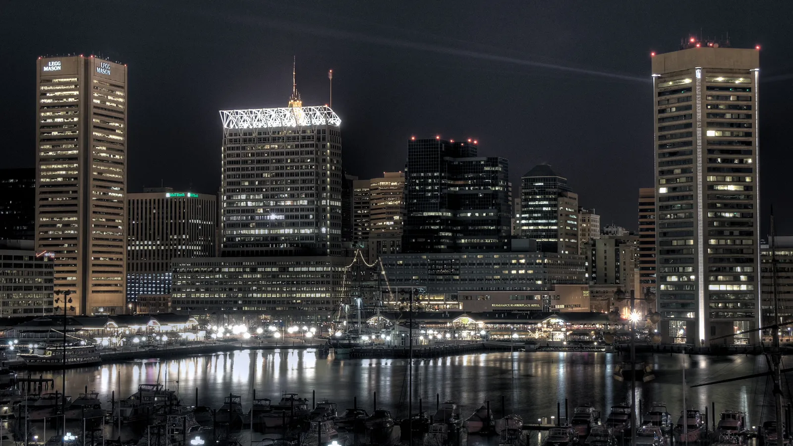 A nighttime cityscape of Baltimore's Inner Harbor with illuminated buildings and boats reflected in the water.