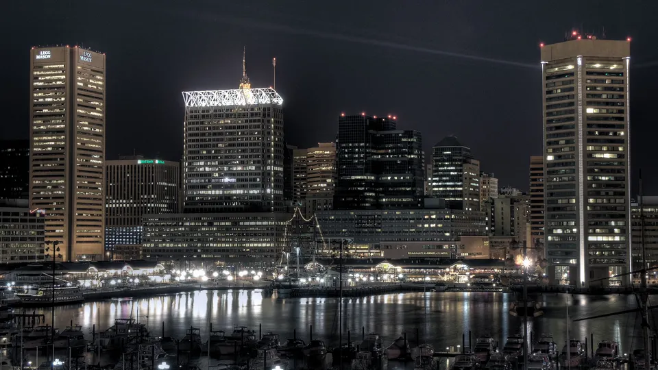 A nighttime cityscape of Baltimore's Inner Harbor with illuminated buildings and boats reflected in the water.