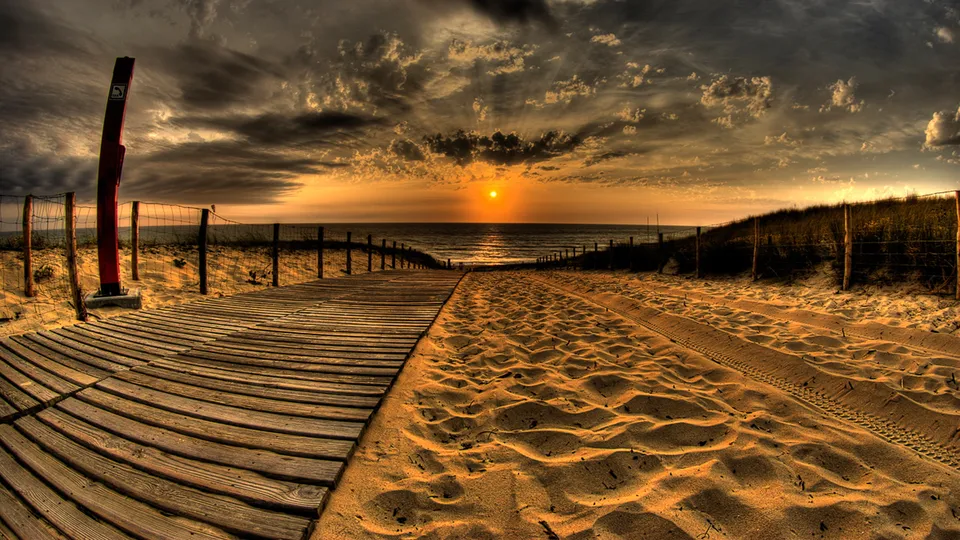A wooden boardwalk leads across sand dunes towards a vibrant sunset over the ocean.