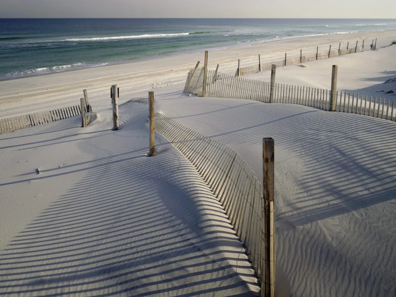 A wooden dune fence casts long, striped shadows across a sandy beach with the ocean in the background.