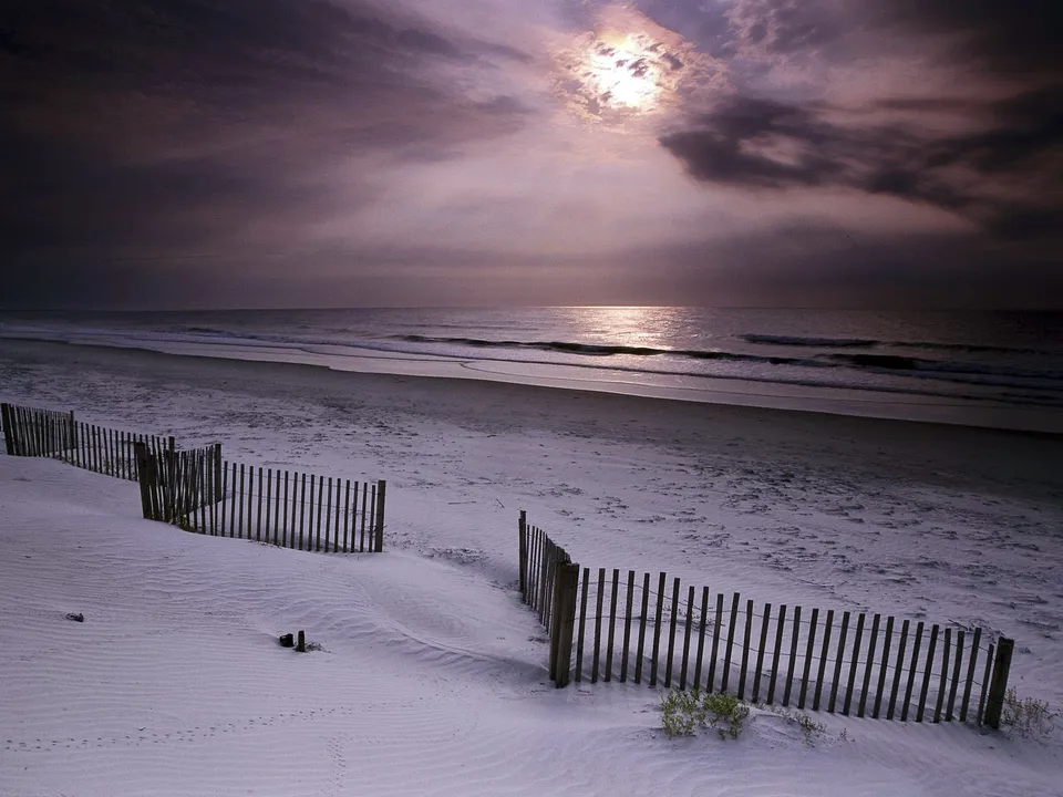 A wooden dune fence lines a sandy beach as the sun sets over the ocean.