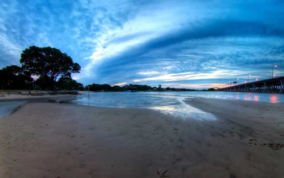 A sandy beach with bird tracks leads to a calm body of water under a dramatic, cloudy sky at dusk, with a bridge in the distance.