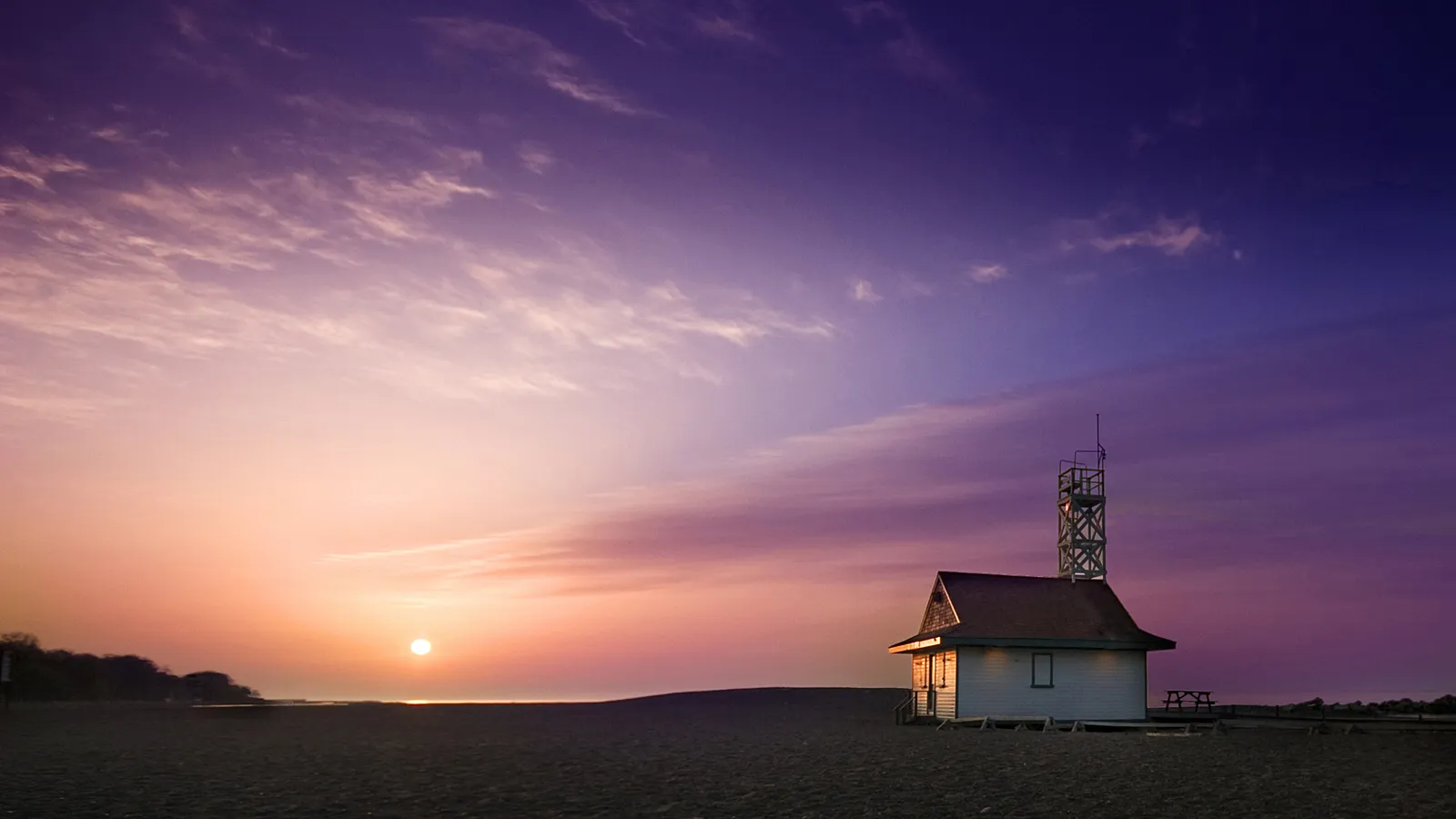 A small beach house with a tower stands on a sandy shore under a vibrant purple and orange sunset sky.