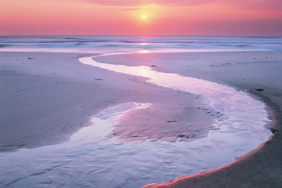 A gentle stream meanders across a sandy beach towards the ocean during a soft pink and orange sunset.