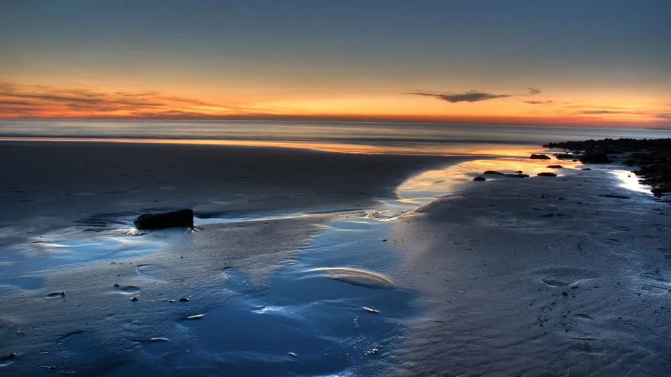 A serene beach scene at sunset with the sky painted in orange and blue hues, reflecting on wet sand.