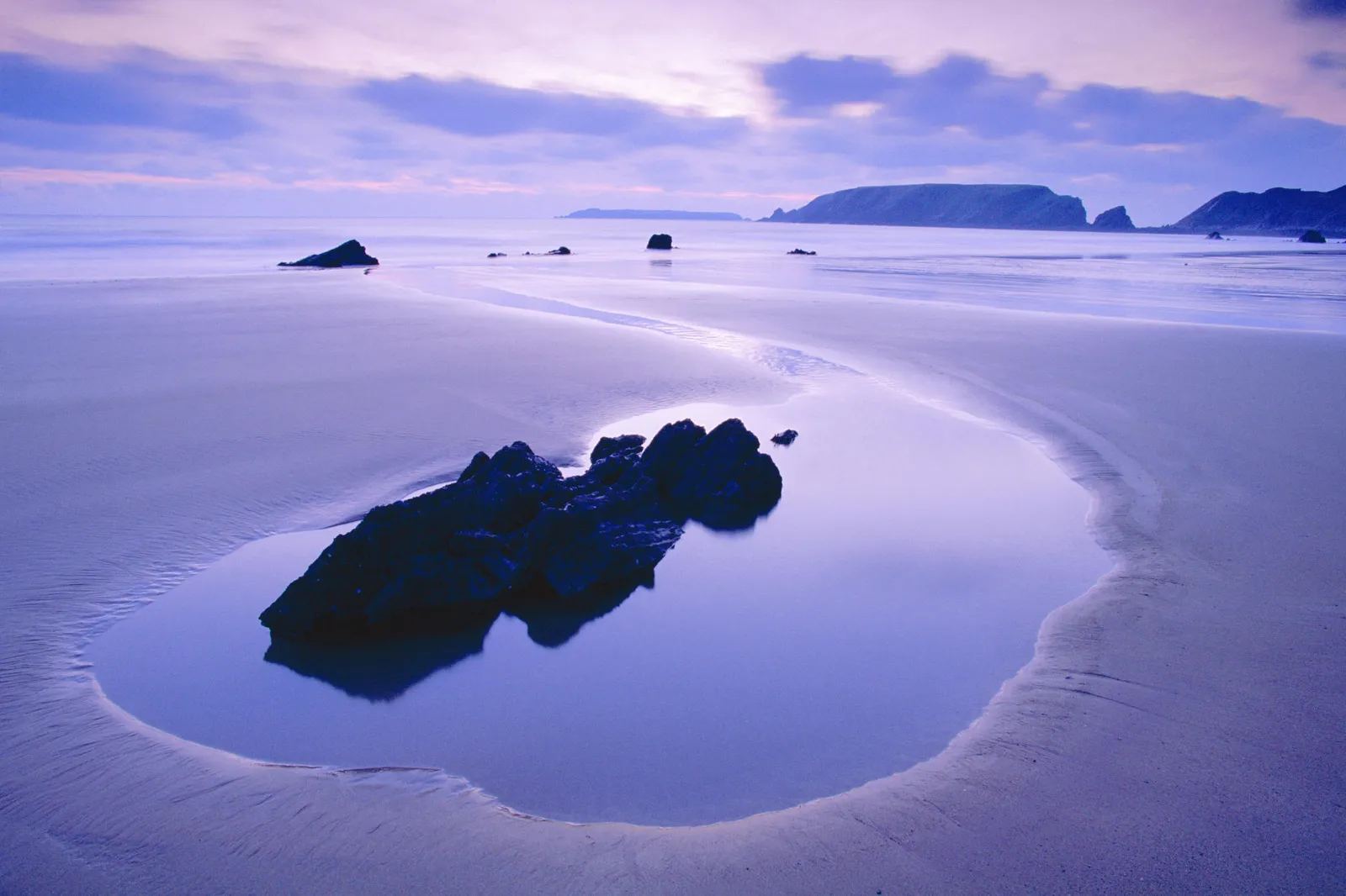 A calm tidal pool on a sandy beach reflects dark rocks and a purple sky at dusk.
