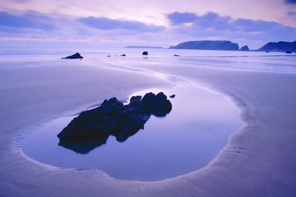 A calm tidal pool on a sandy beach reflects dark rocks and a purple sky at dusk.