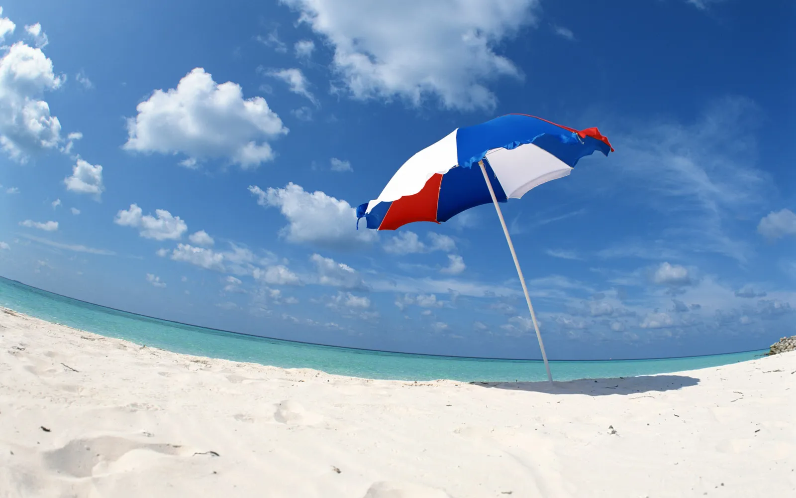 A red, white, and blue beach umbrella stands on a white sand beach with turquoise water and a blue sky with clouds.