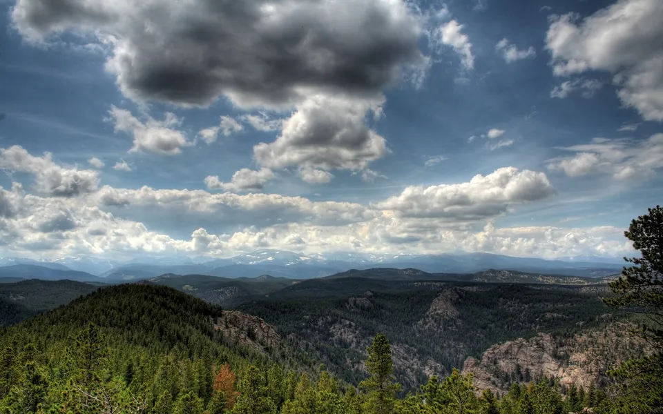 A wide vista of evergreen forests under a dramatic sky with large, billowing clouds.