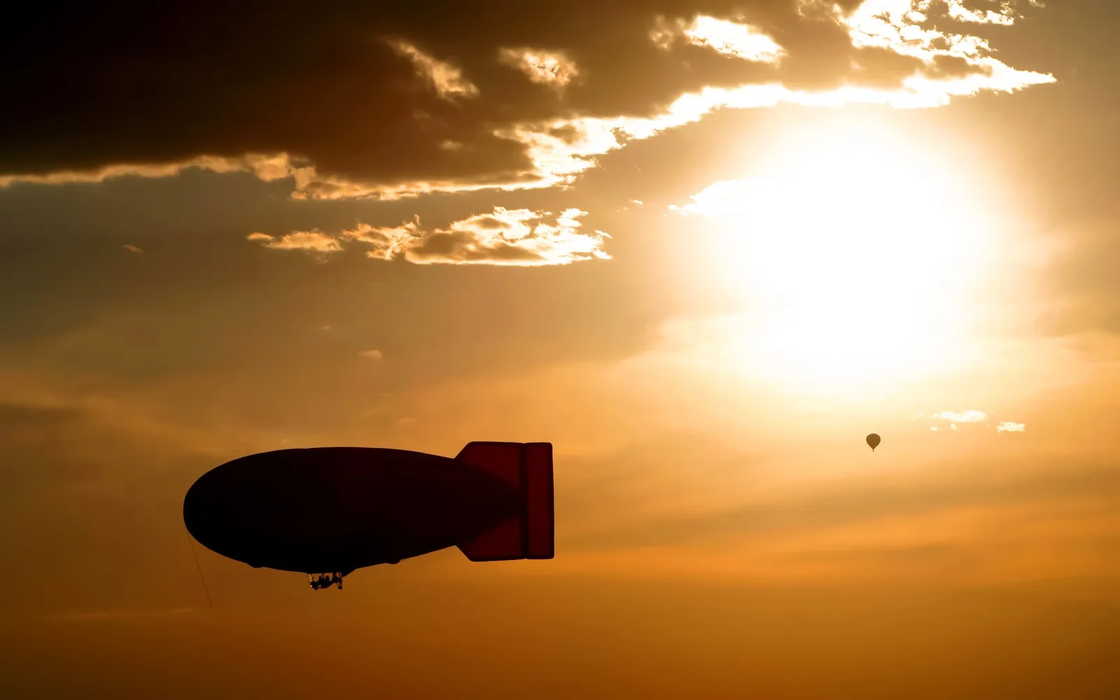 A large blimp and a small hot air balloon are silhouetted against a bright, cloudy sunset sky.