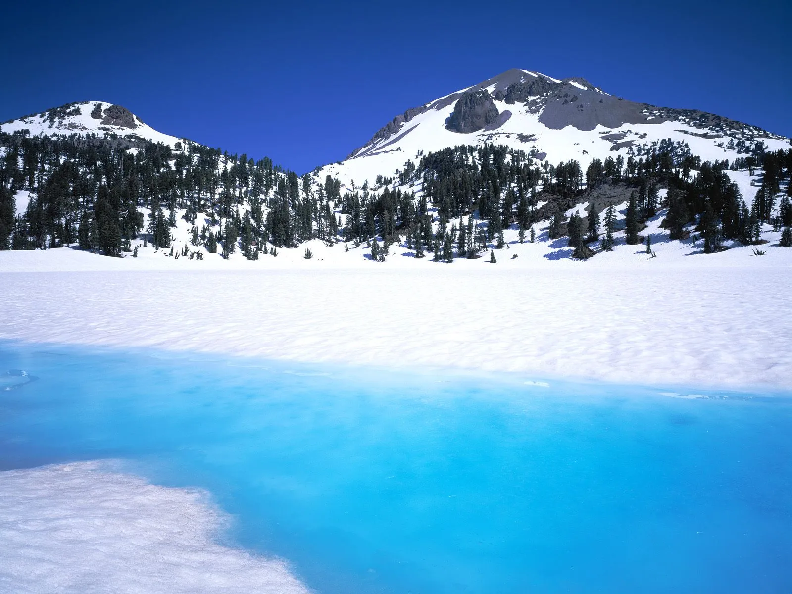 A bright blue, partially frozen lake in the foreground with snow-covered mountains and pine trees in the background under a clear blue sky.