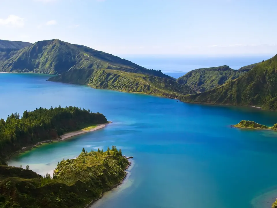 A wide view of a bright blue crater lake surrounded by lush green, rolling hills under a clear sky.