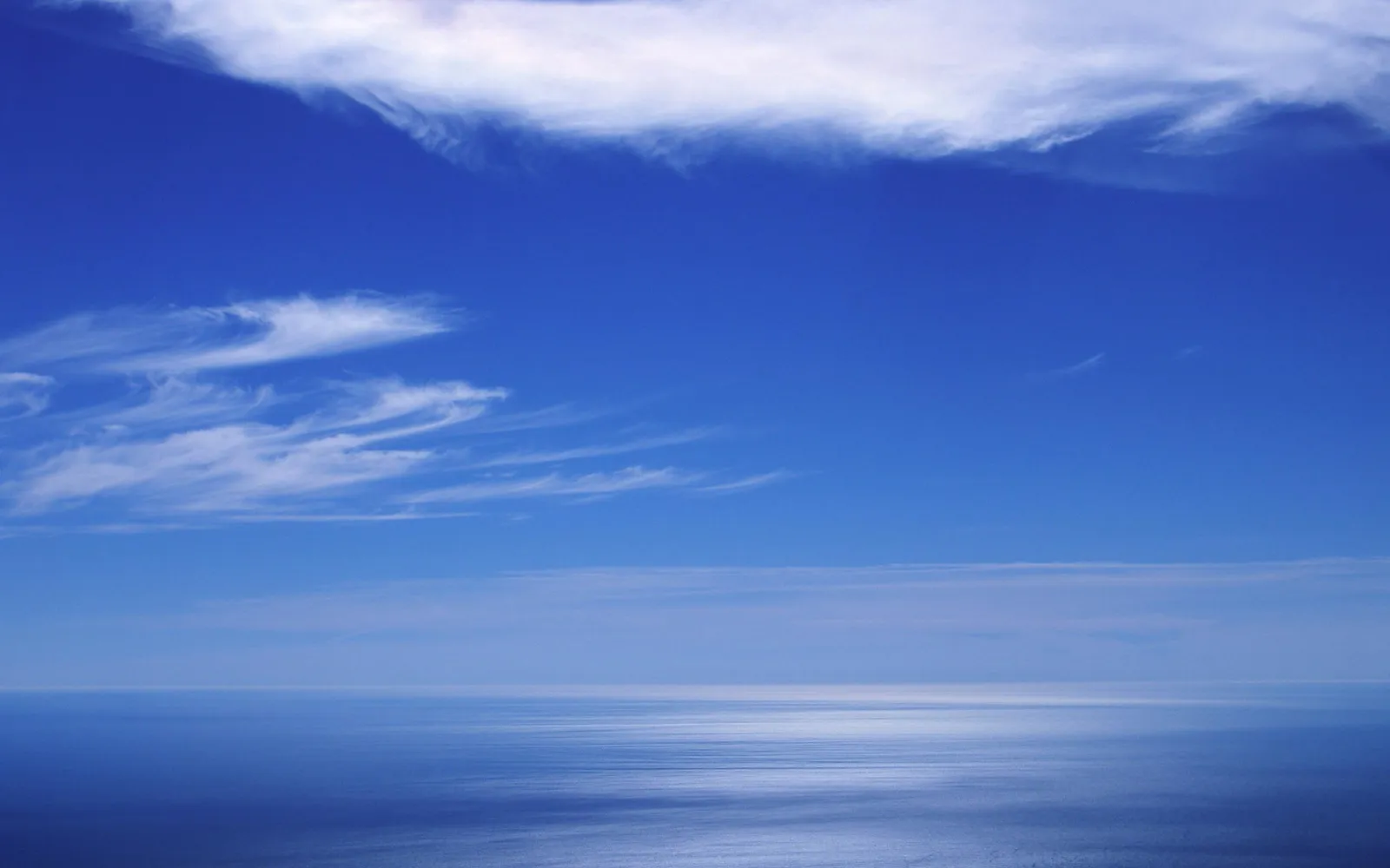 A wide expanse of deep blue sky with wispy white clouds above a calm, reflective ocean.
