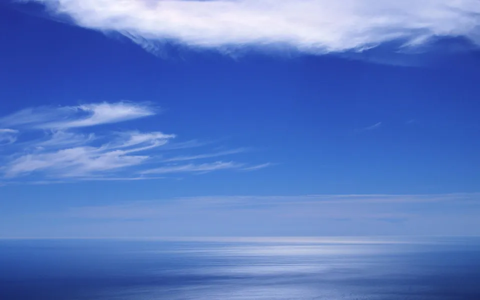 A wide expanse of deep blue sky with wispy white clouds above a calm, reflective ocean.