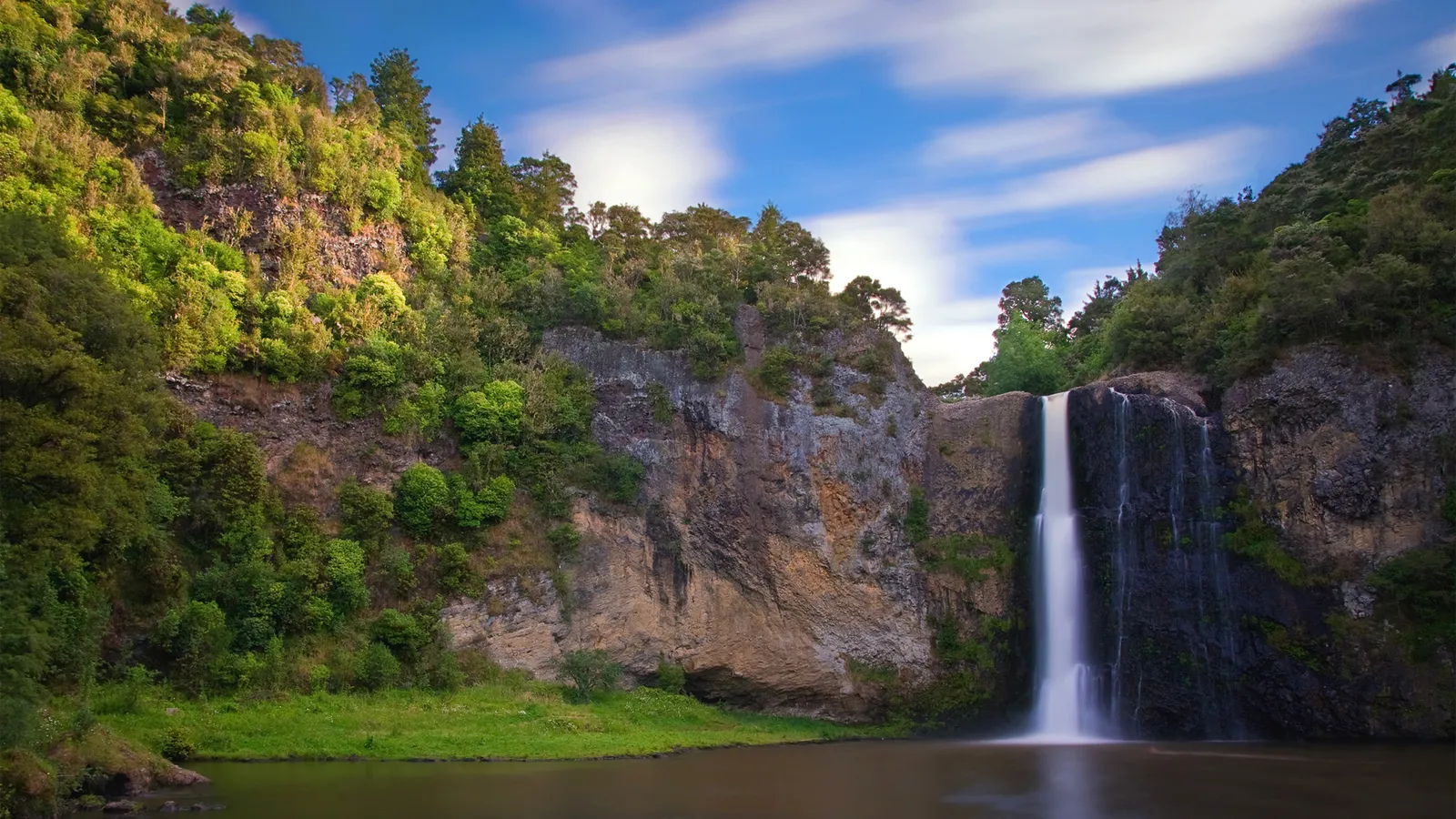 A tall waterfall cascades down lush green cliffs under a blue sky with streaky clouds.