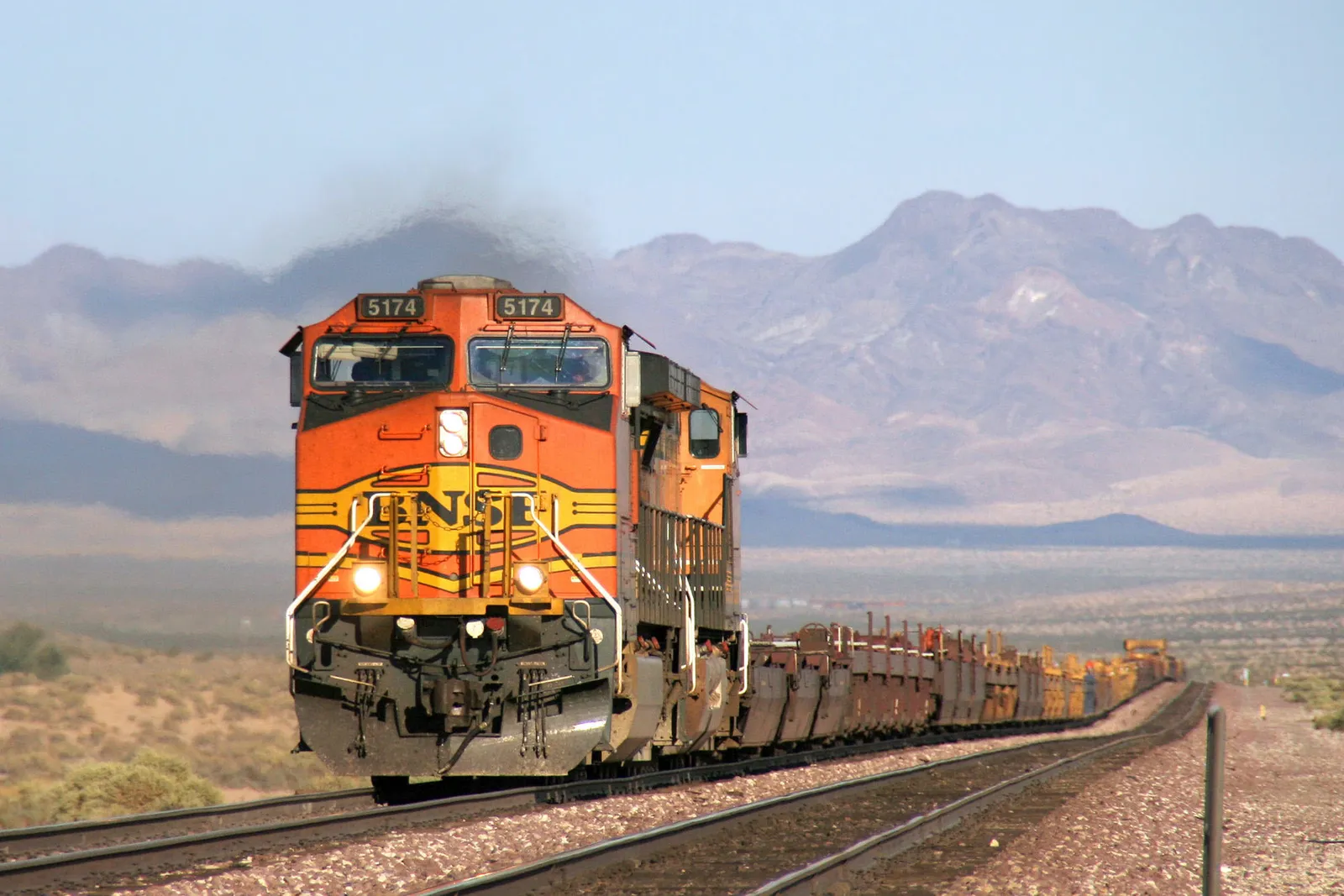A BNSF freight train with an orange and yellow locomotive travels on railroad tracks through a desert landscape with mountains in the background.