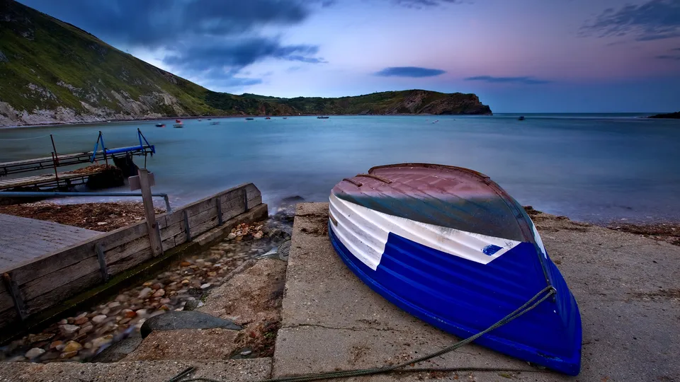 A blue and white boat rests on a concrete shore next to a calm bay and green hills.