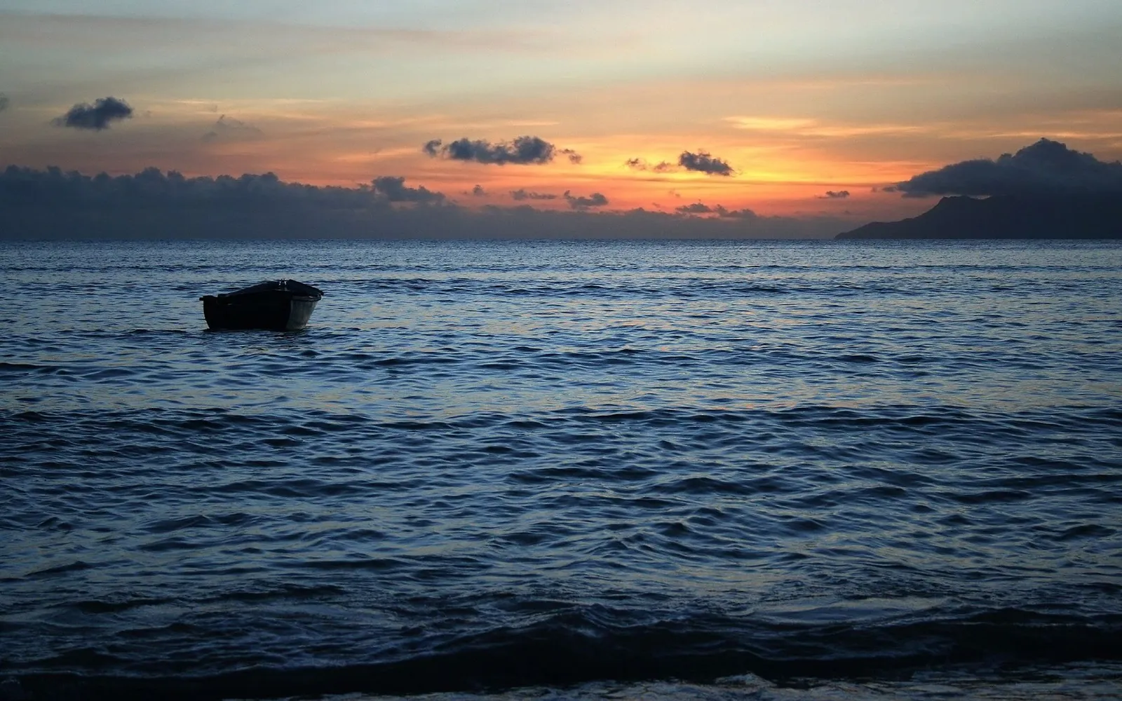 A lone rowboat floats on the ocean during a colorful sunset with clouds and a distant island.