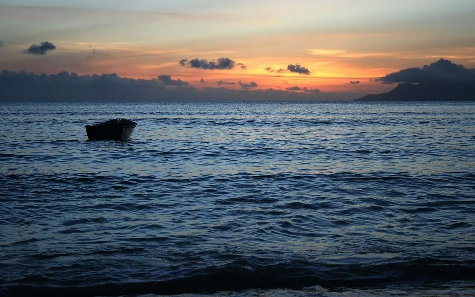 A lone rowboat floats on the ocean during a colorful sunset with clouds and a distant island.