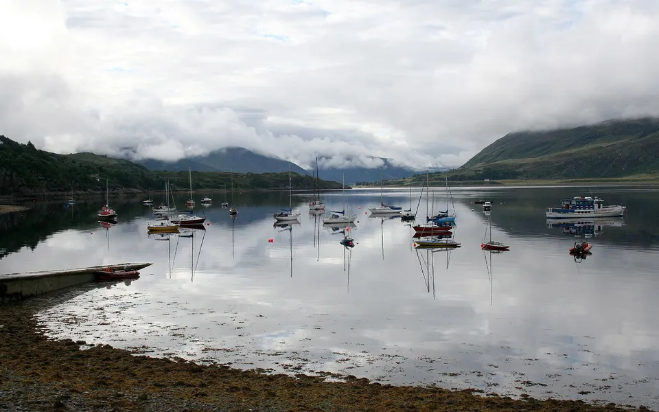 A calm lake with many sailboats and a ferry, reflecting the cloudy sky and misty mountains.