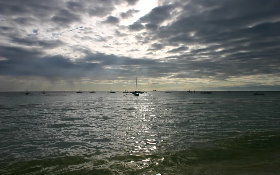Several small fishing boats are anchored in the ocean under a dramatic sky filled with dark clouds and sunbeams.
