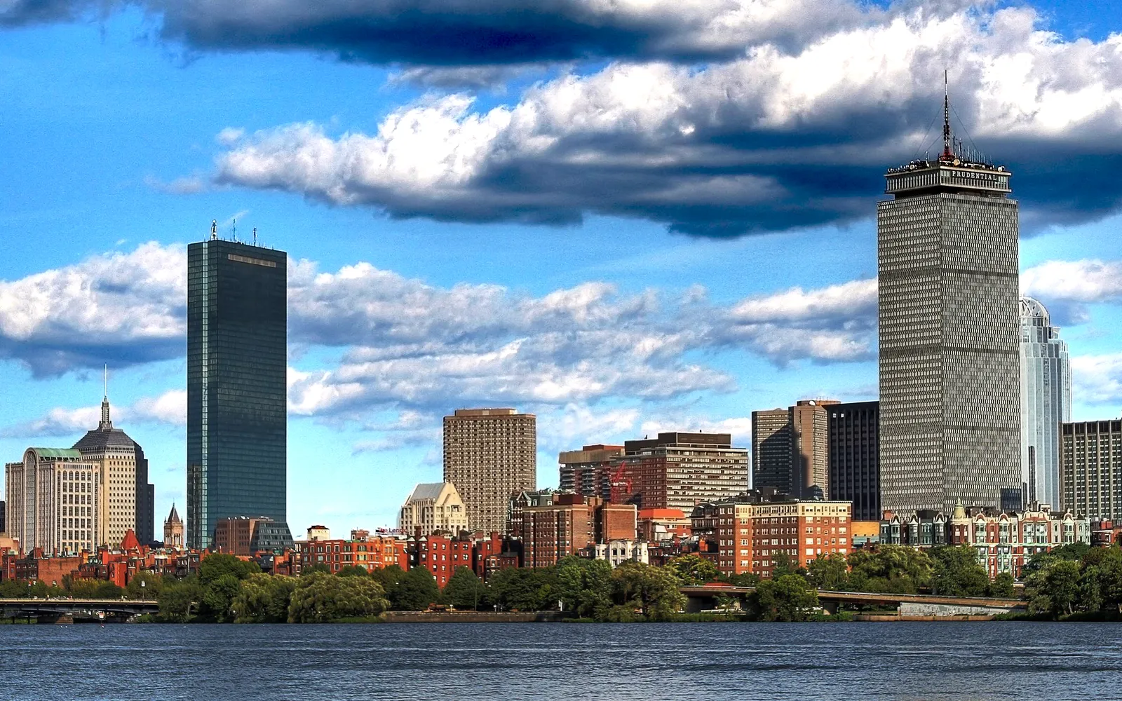A wide view of the Boston skyline with tall buildings reflected in the water under a dramatic sky.