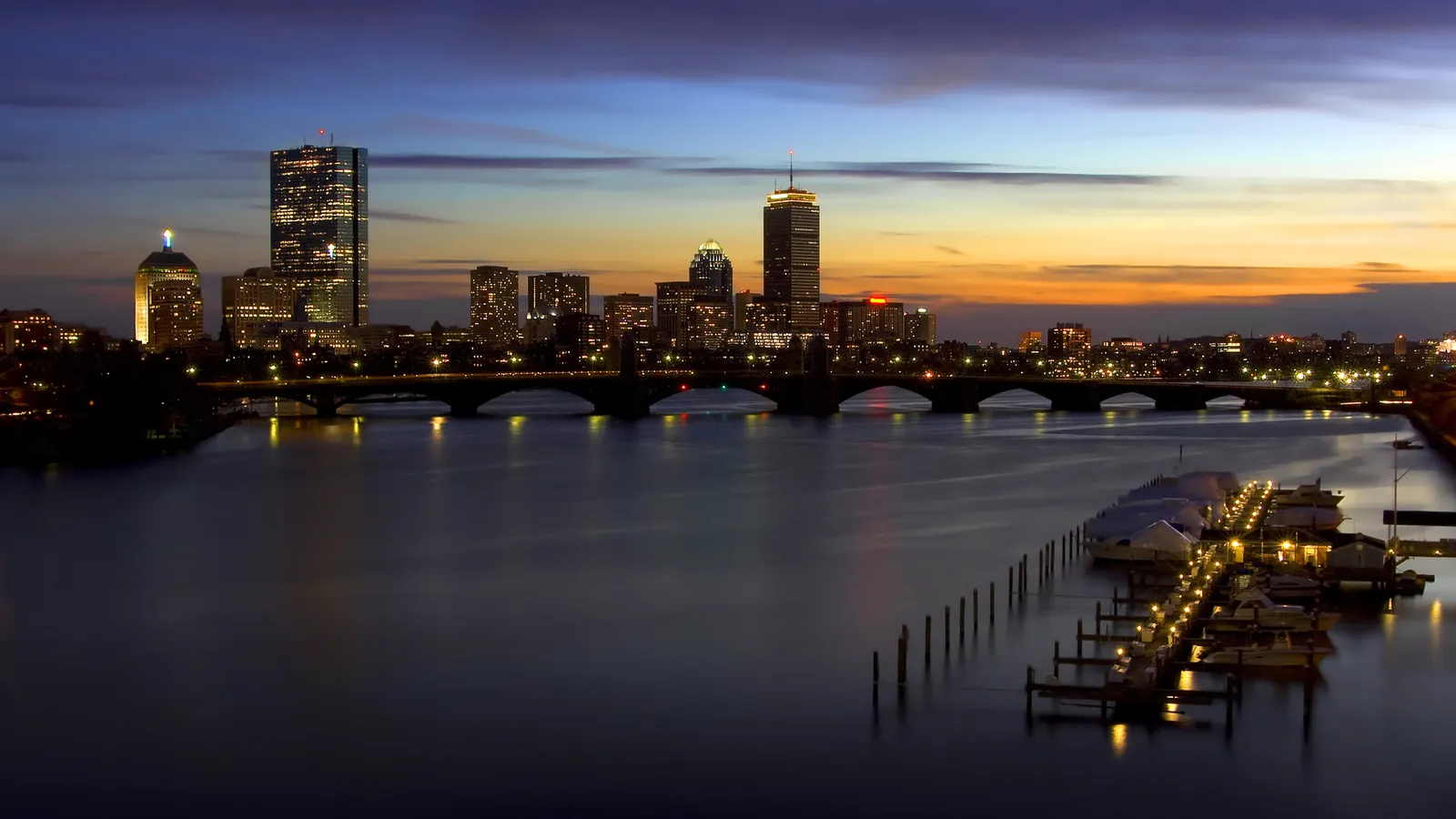 A cityscape of Boston at dusk with illuminated buildings and a bridge over a calm river.